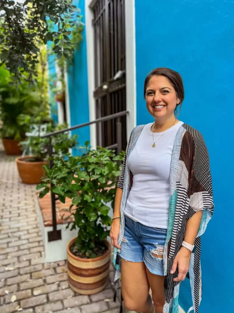 Woman standing in front of blue wall with jean shorts, white shirt and multi colored cover up.
