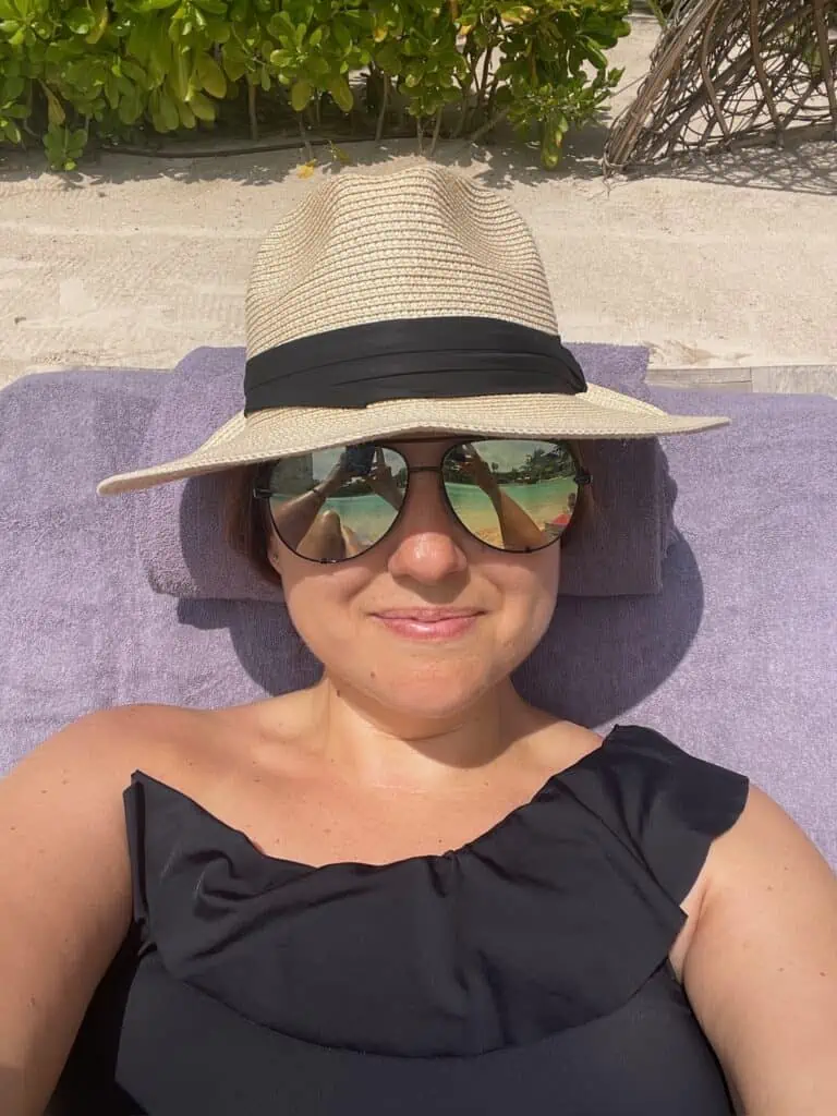 Woman laying at the beach with black bathing suit on. She is wearing a sun hat and sun glasses.