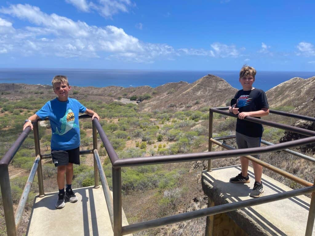 Two young boys standing on lookout at the top of Diamond Head. Mountains and Ocean in background. 