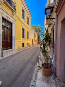 Empty street in Athens with yellow and pink buildings and potted plant on sidewalk.