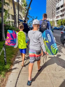 2 young boys with dad walking on sidewalk in Waikiki holding boogie boards.